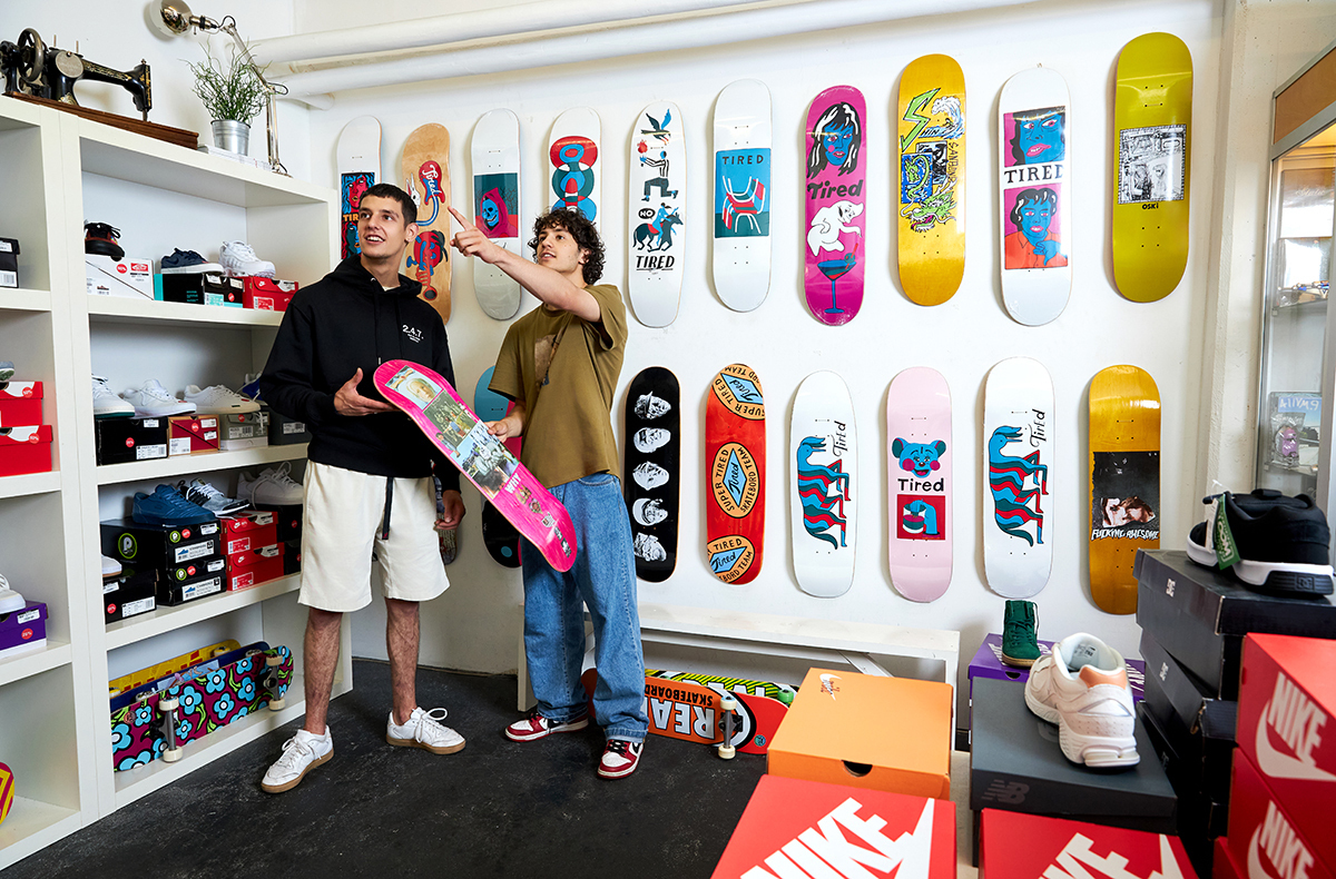Twee jonge mannen staan in een winkel met skateboarddecks aan de muur. Eén houdt een skateboard vast, de ander wijst naar een deck.