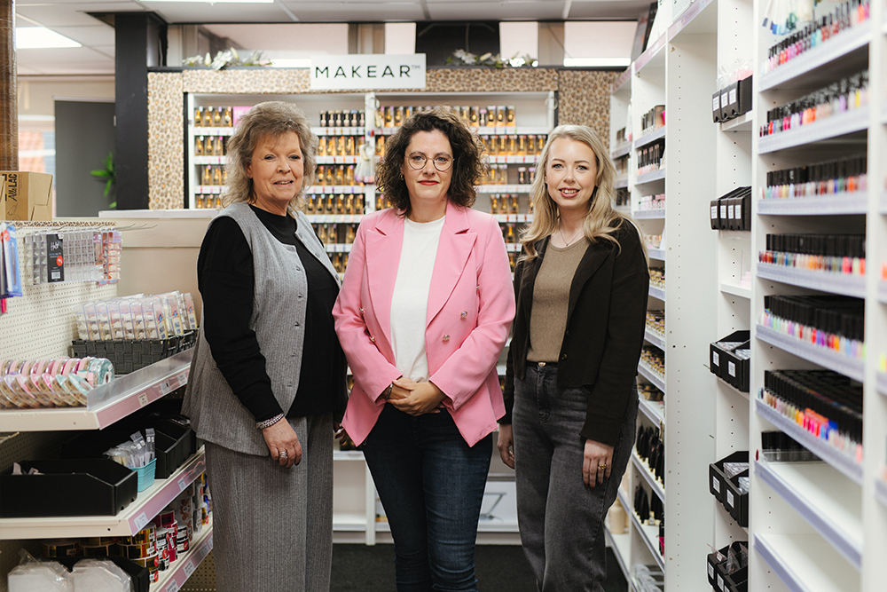 Drie vrouwen staan naast elkaar in een winkel met nagelverzorgingsproducten op de achtergrond.