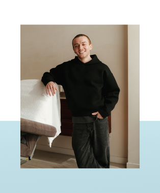 Een lachende man in een zwarte hoodie leunt met een arm op een stoel in een kamer.