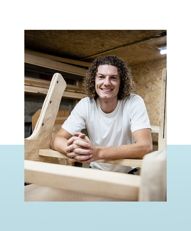 Lachende man met krullend haar leunt op een houten werkbank in een houtwerkplaats.