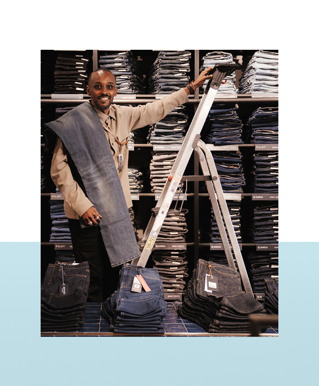 Man staat voor een wand met opgestapelde spijkerbroeken en een ladder.