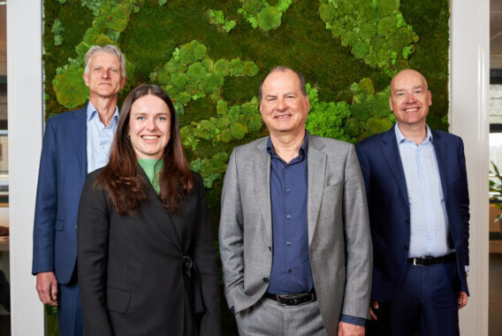 Vier personen staan naast elkaar in een kantoorruimte met een groene wand op de achtergrond.