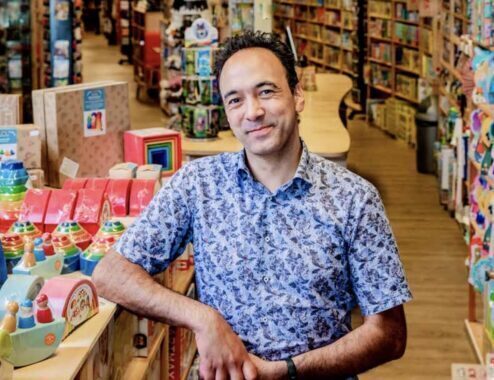 Man leunt tegen een tafel in een winkel met speelgoed en boeken.