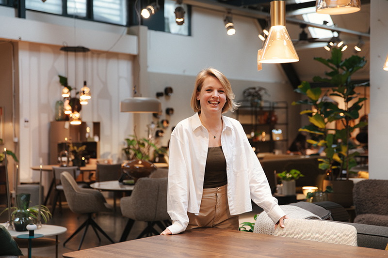 Vrouw staat lachend achter een houten toonbank in een winkel met planten op de achtergrond.