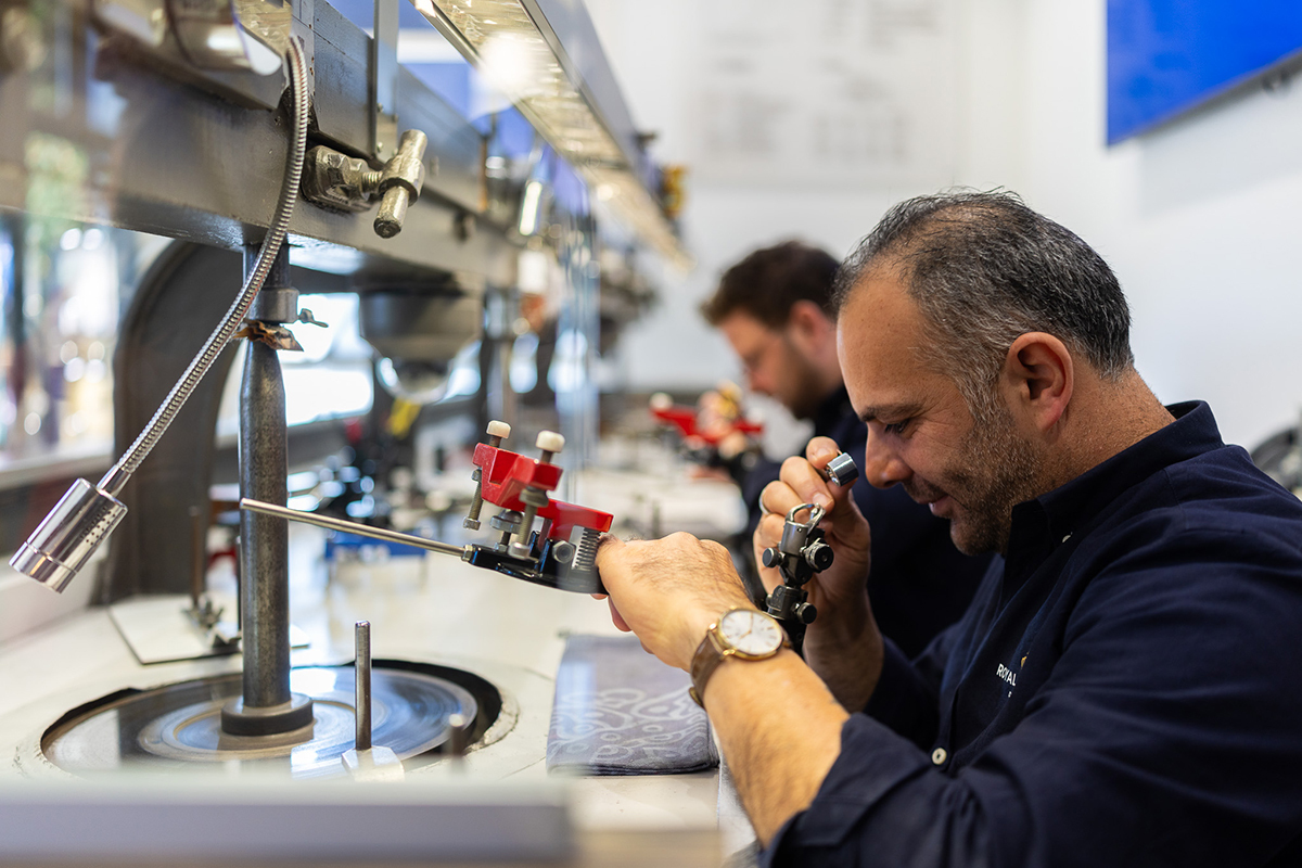 Man met baard werkt geconcentreerd aan een machine in een werkplaats, met een collega op de achtergrond.