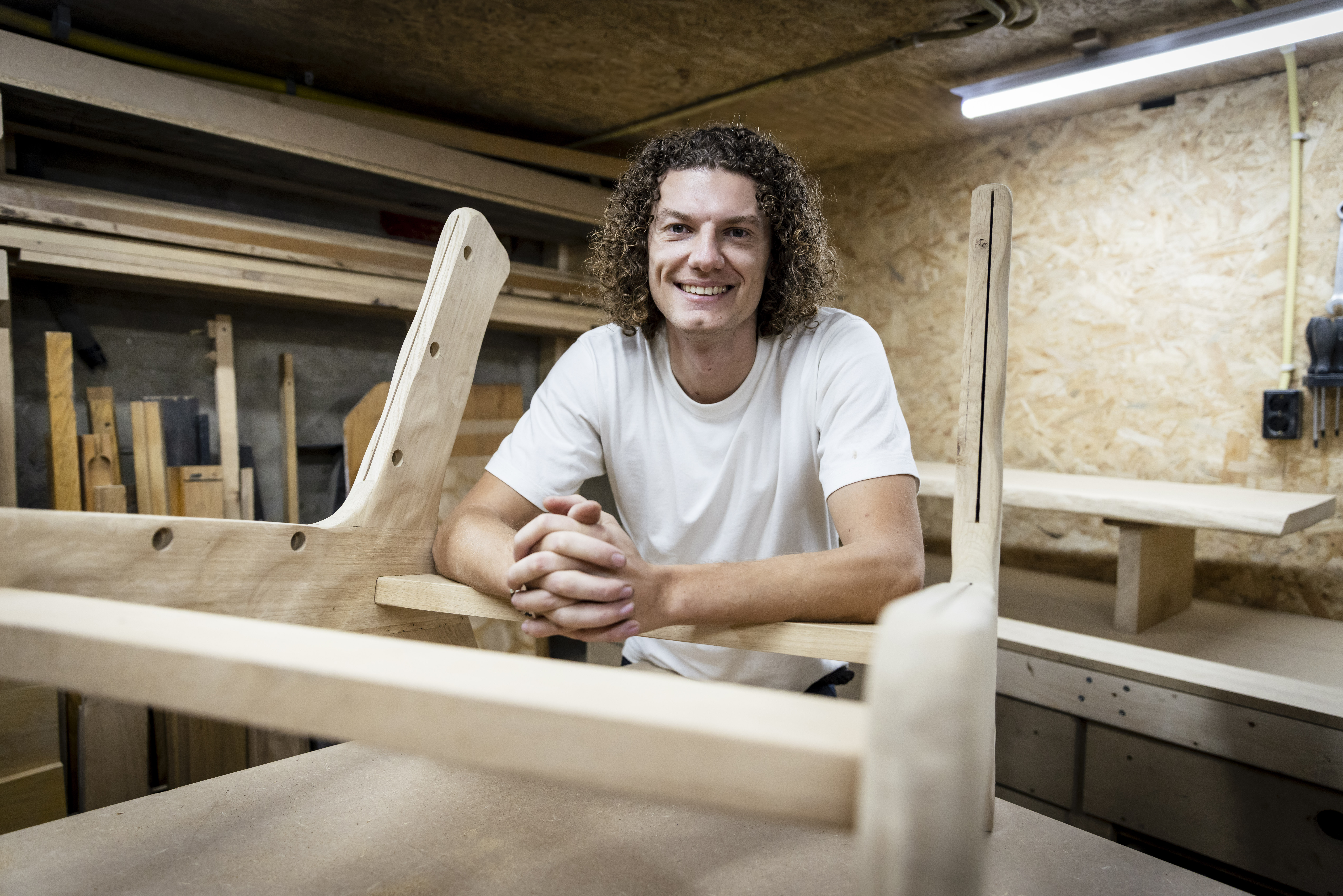 Lachende man met krullend haar leunt op een houten werkbank in een houtwerkplaats.