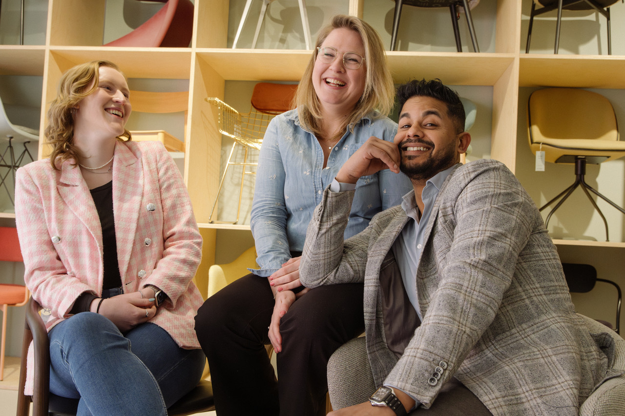 Drie personen zitten lachend tussen stoelen in een meubelwinkel.