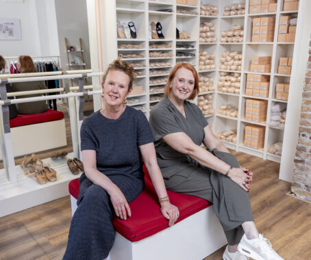 Twee vrouwen zitten naast elkaar in een balletwinkel met schoenen in schappen achter hen.