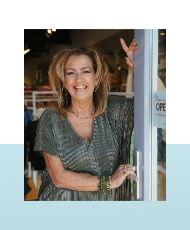 Vrouw staat lachend in de deuropening van een kledingwinkel.