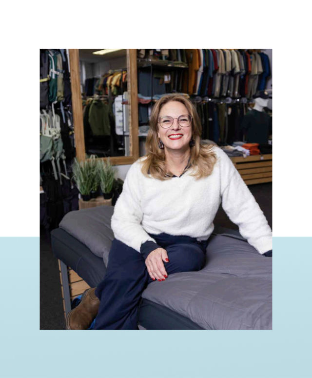 Vrouw met bril zit lachend in een kledingwinkel met kledingrekken op de achtergrond.