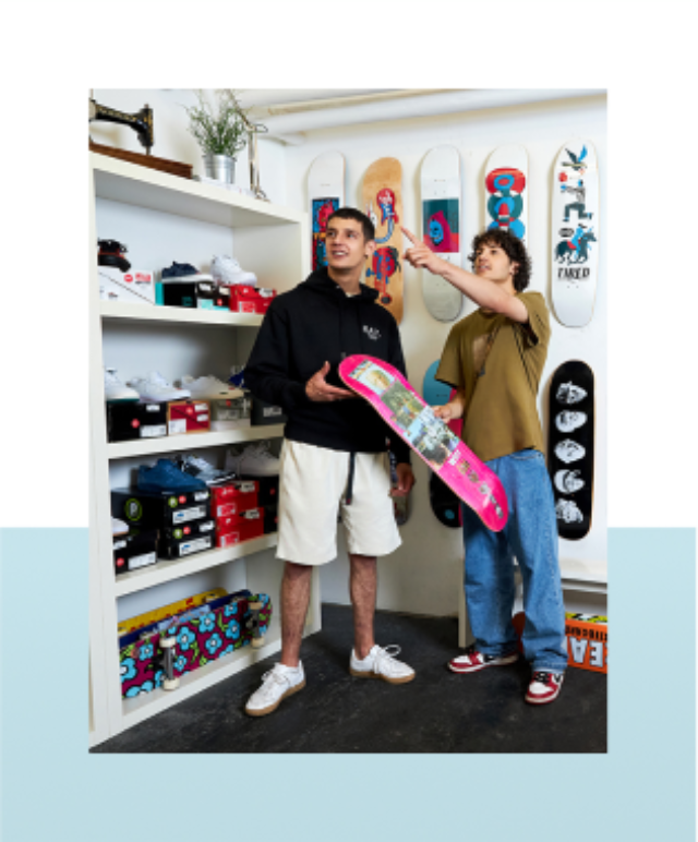 Twee personen staan in een winkel met skateboards; één houdt een skateboard vast terwijl de ander wijst.