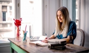 Vrouw zit aan een houten tafel bij een raam en schrijft in een notitieboek. Op tafel liggen papieren en een laptop.