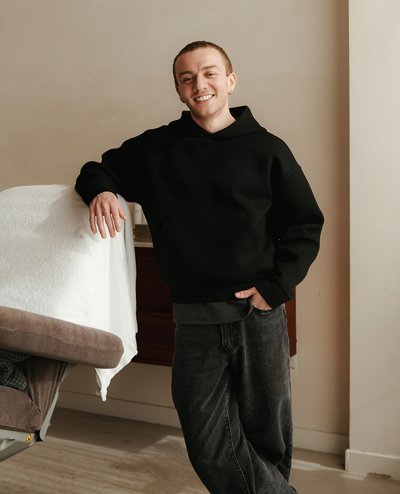 Een lachende man in een zwarte hoodie leunt met een arm op een stoel in een kamer.