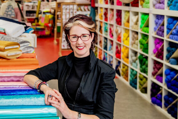 Vrouw staat in een winkel met gekleurde bollen wol in vakken achter haar.