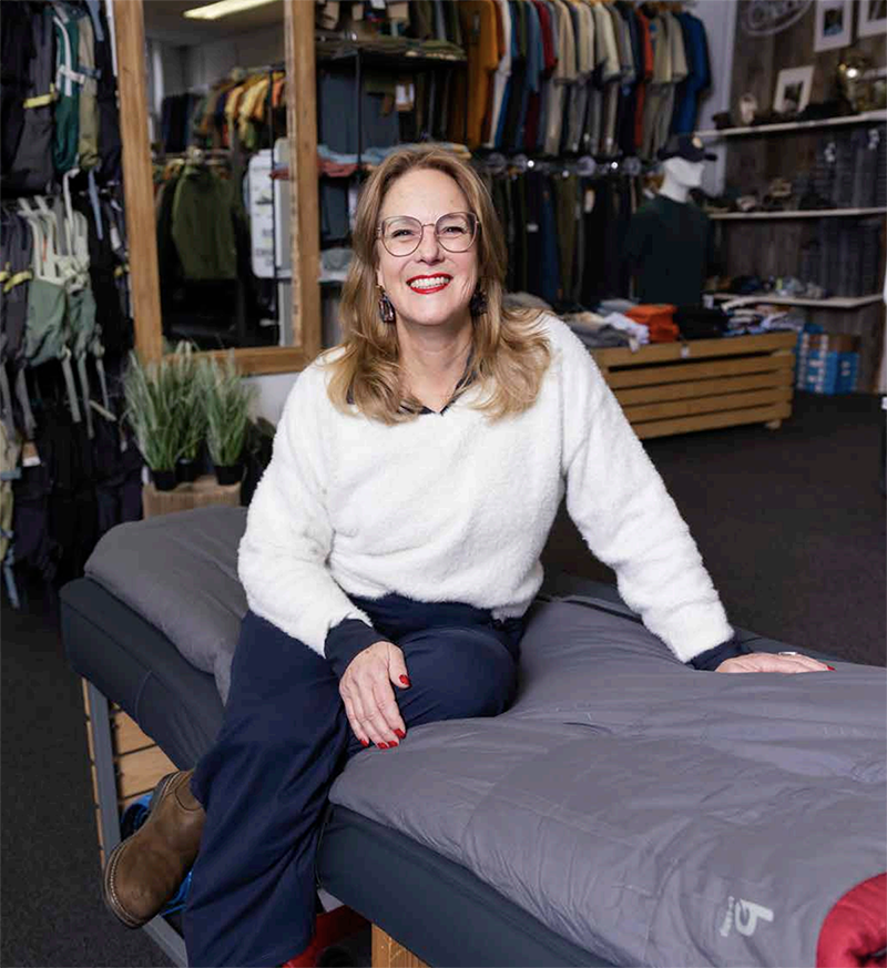 Vrouw met bril zit lachend in een kledingwinkel met kledingrekken op de achtergrond.