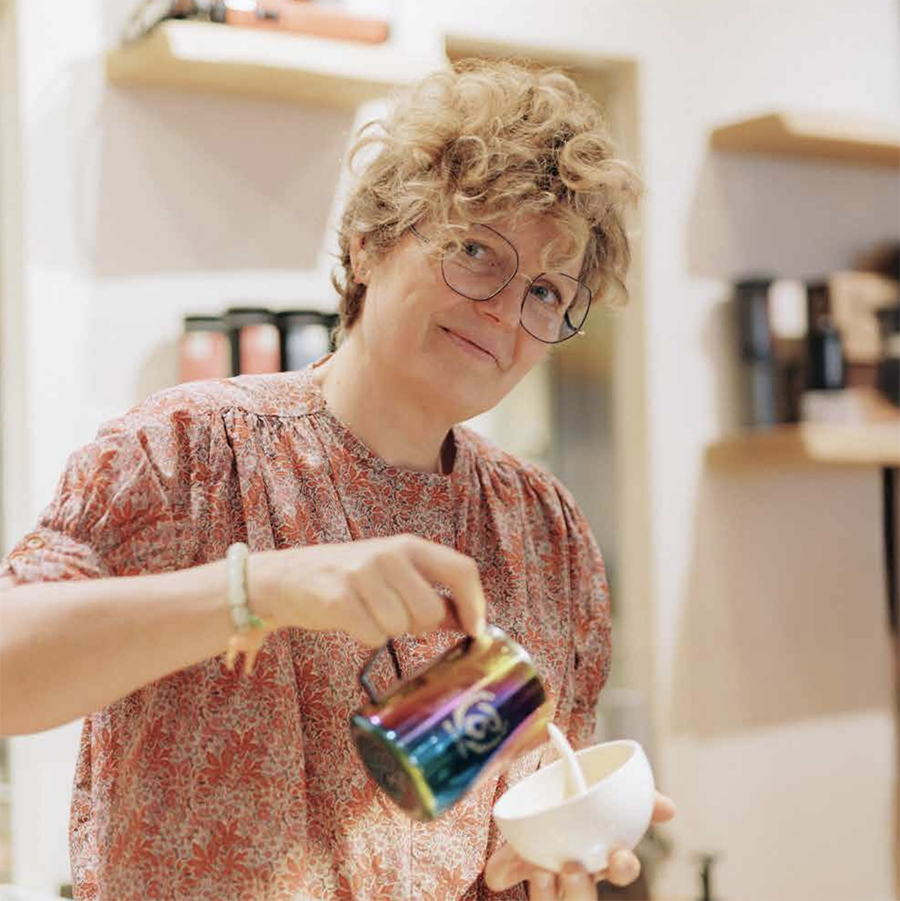 Vrouw met bril en krullend haar schenkt iets uit een kleurrijk pak in een kopje in een winkel met gevulde schappen op de achtergrond.