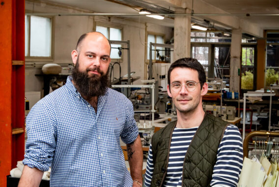 Twee mannen staan naast elkaar in een werkplaats met machines op de achtergrond.
